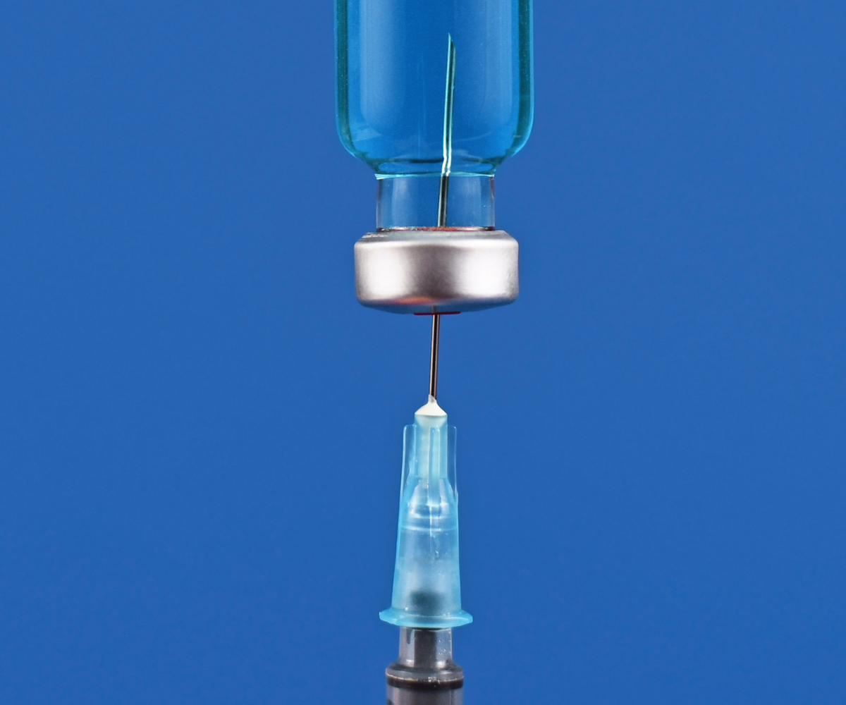 A needle is held up and pierces the rubber stopper of a glass bimix medication vial.